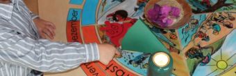 Zu sehen ist die Hand eines Kindes. Das Kind zeigt auf ein Bild auf dem Tisch. Dieses gehört zu einer bildlichen, im Kreis angeordneten Darstellung der Jahreszeiten. 