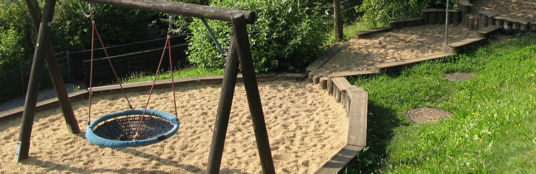 Auf der linken Bildseite ist eine Korbschaukel in einem Sandkasten zu sehen. Der Sandkasten ist mit zwei weiteren Sandkästen verbunden, welche stufenförmig angelegt sind. Des weiteren sieht man Wiese und Büsche.