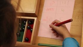 Zu sehen ist ein Mädchen, dass Zahlen und Strichblöcke schreibt. Auf der linken Seite steht eine Shcachtel mit Murmeln. 
