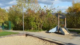 Zu sehen ist ein Teil eines Sandkastens. Ein Weg aus Platten trennt den Sandkasten von einer Rutsche mit Treppe für Kleinkinder. Im Hintergrund sind viele Büsche und Sträucher. 